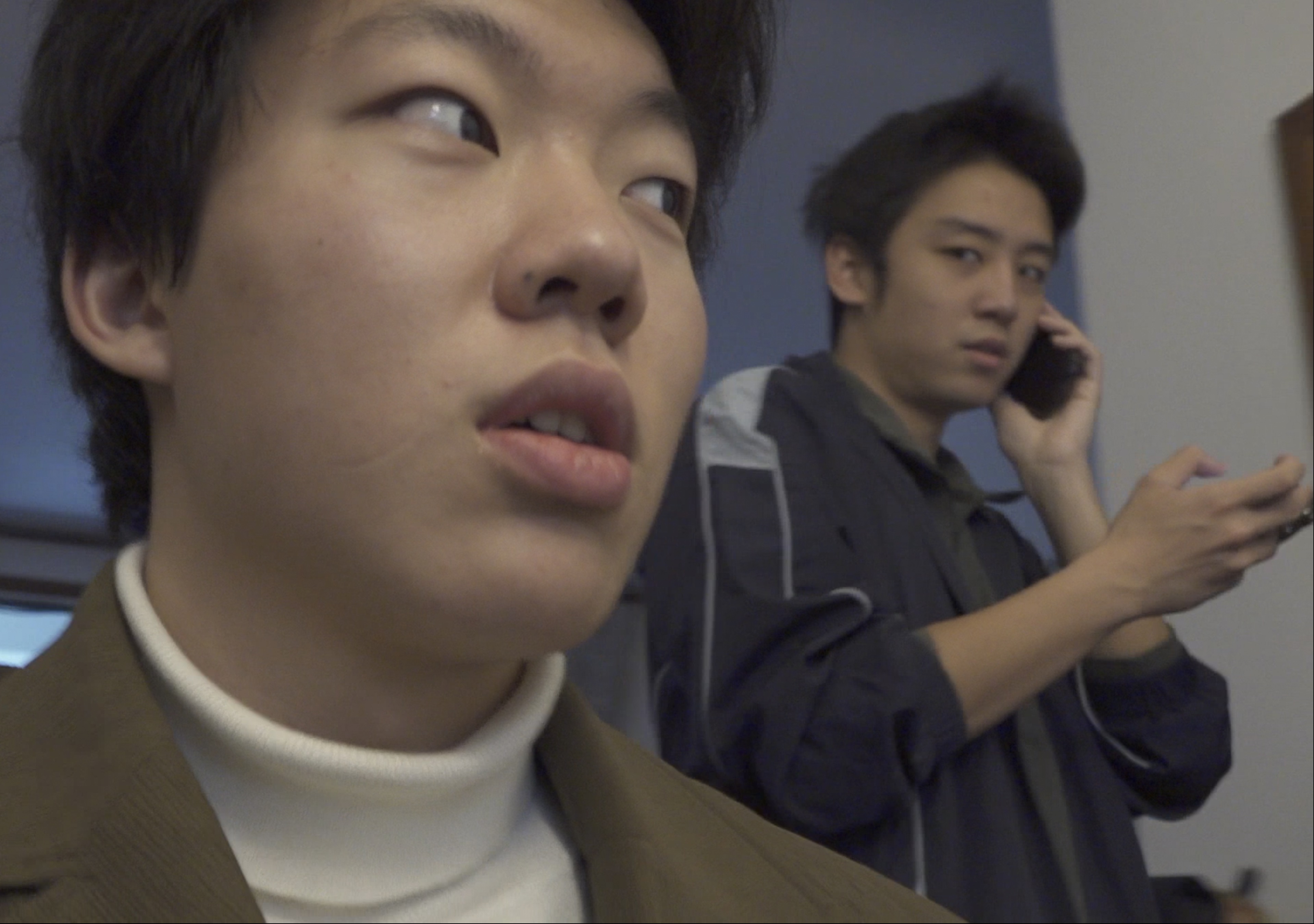 A still from Tutor’s Diary, with one character in the foreground and another speaking on the phone behind him.